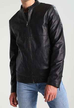 Man wearing a black leather zip-up jacket with stand collar and light blue jeans against a plain light background.
