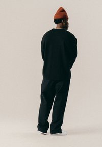 Man met een roestkleurige beanie, een oversized zwarte trui, zwarte broek en witte sneakers, staand en zijwaarts gekeerd tegen een effen achtergrond.