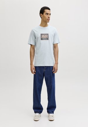 Muž stojí, má na sebe svetlomodré tričko Jack & Jones, voľné tmavomodré džínsy a svetlé topánky, pozerá sa doprava na jednoduchom pozadí.