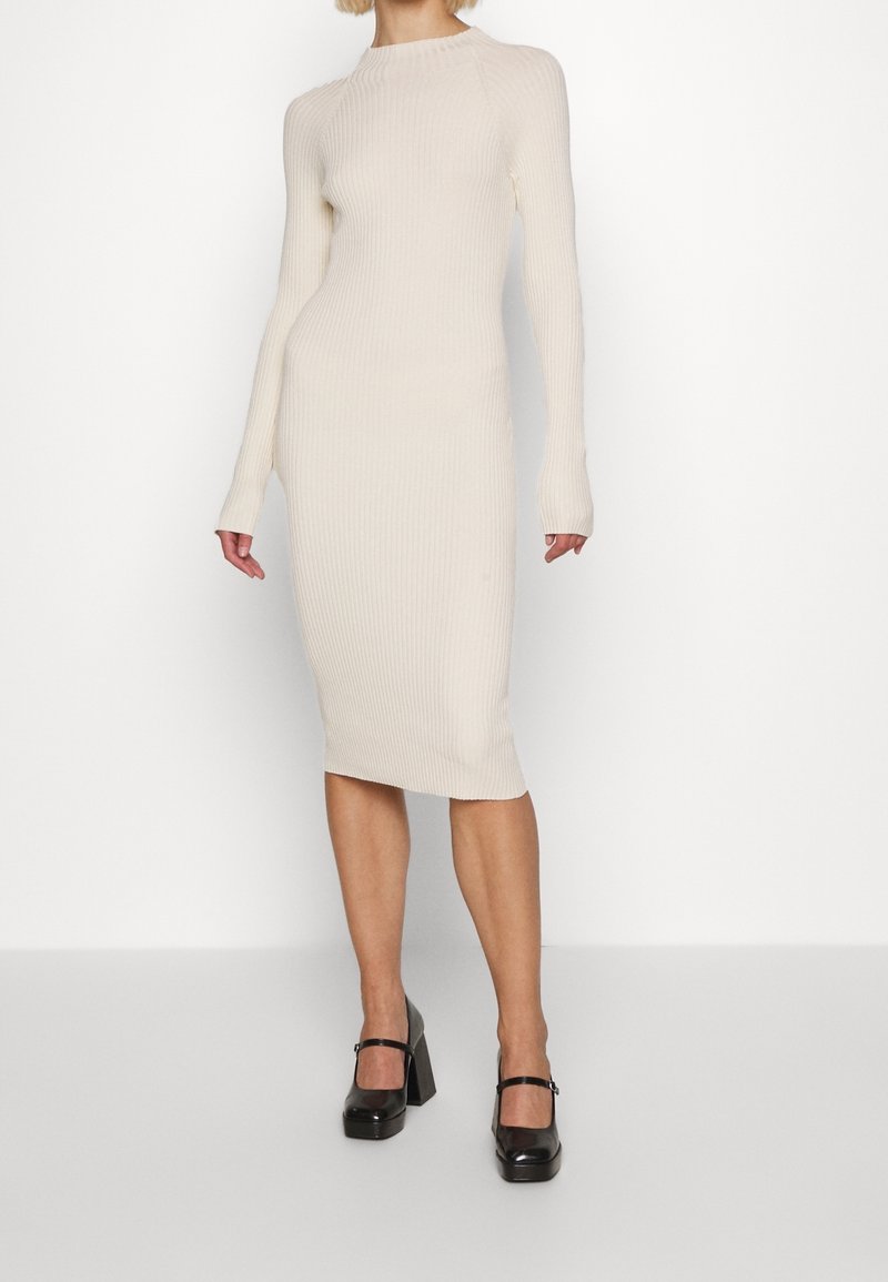 Femme portant une robe midi ajustée à manches longues en maille côtelée couleur crème, associée à des chaussures Mary Jane noires à talons épais, debout sur un fond blanc.