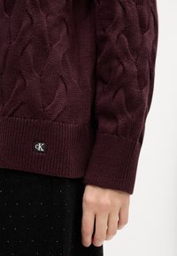 Burgundi kabelių mezgimo megztinis su giliomis rankovėmis, su maža juoda cK logotipo etikete prie apdailos. Medžiaga atrodo minkšta ir tekstūruota.