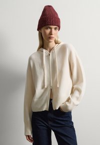 Woolrich zip-up bianco sporco con cappuccio, polsi e orlo a costine; abbinato a un berretto di maglia color amaranto. Tessuto leggero con una consistenza morbida.