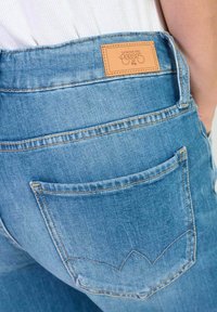 Un jean en denim bleu clair présente une étiquette en cuir sur la taille et un design distinct de poche arrière avec des coutures décoratives.