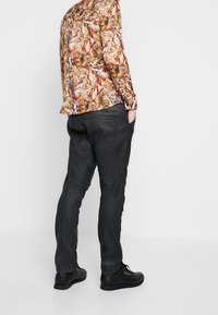Pantalones negros de corte ajustado con detalles de costura, combinados con una camisa de manga larga de patrón colorido. Tiene bolsillos laterales y zapatos negros.