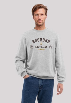 Gray sweatshirt with crew neck featuring printed text and anchor design in dark brown and mustard yellow. Paired with blue denim jeans.