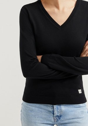 Personne portant un pull noir en V, les bras croisés et un jean bleu clair sur un fond uni clair.