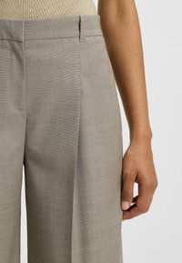 Pantaloni beige su misura con un tessuto strutturato, gambe larghe e pince frontali. La cintura è liscia e le tasche laterali sono integrate.