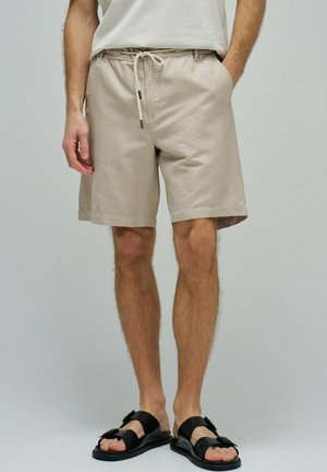 Hombre con pantalones cortos beige con cordón, camiseta blanca y sandalias negras con hebilla, de pie con las manos en los bolsillos.