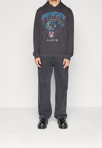 Huppari harmaalla hupulla, jossa on pantterigrafiikka ja "TOMMY"-teksti, yhdistettynä leveälahkeisiin harmaisiin farkkuihin ja mustiin kenkiin. Yksinkertainen tausta.