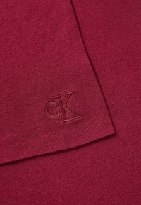 Tejido de punto burdeos que exhibe una textura suave con un detalle de logo en relieve "cK", mostrando un diseño simple y elegante.