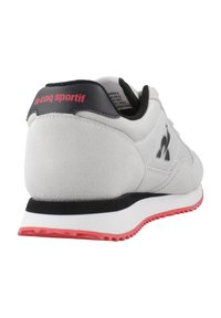 Zapatilla de deporte gris claro con textura de ante y acentos en negro. Presenta una suela de goma roja y un logo en el talón. Diseño atlético clásico.