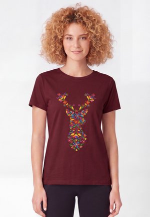 Frau mit lockigem Haar, die ein dunkelrotes T-Shirt mit einem bunten, floralen Hirschkopf-Design auf der Vorderseite trägt.