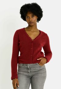 Cárdigan rojo hecho de material suave con cuello en V, bordes festoneados y cierre de botones, combinado con vaqueros grises de talle alto.