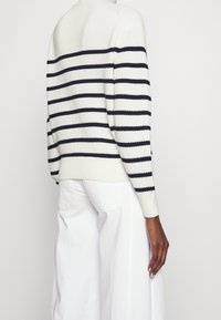 Persona vistiendo un suéter de punto blanco con rayas negras horizontales y pantalones blancos, de pie contra un fondo claro y liso.