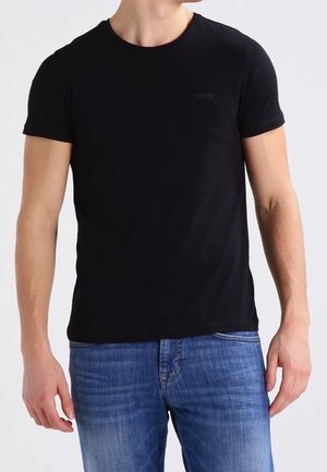 Mężczyzna ubrany w dopasowaną czarną koszulkę z krótkim rękawem i niebieskie dżinsy, stojący przodem z rękami swobodnie opuszczonymi wzdłuż ciała.