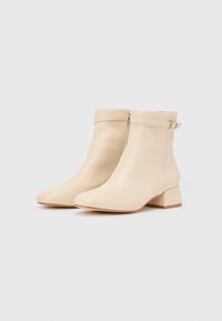 Bottines en cuir beige avec un bout légèrement pointu et un talon épais. Elles disposent d'une fermeture éclair et d'une sangle décorative avec une boucle dorée.