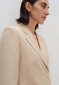 Blazer beige con colletto a punta, chiusura con un bottone, tessuto testurizzato e silhouette strutturata. Orecchino a cerchio semplice in argento visibile.