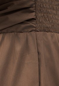 Καφέ ύφασμα με πιετά και σφηκοφωλιασμένη υφή, που παρουσιάζει λεπτομερείς ραφές και πτυχές σε ένα ένδυμα.