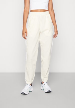 Femme portant un pantalon de survêtement couleur crème avec taille et poignets élastiques, associé à des chaussures de sport blanches et un haut court blanc.