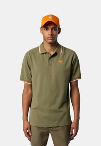 Polo shirt verde oliva con colletto a righe arancioni e bianche, con un piccolo logo arancione. Abbinato a un cappellino arancione.