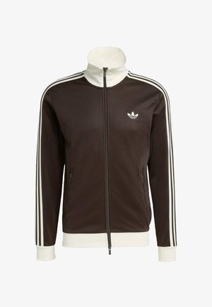 Smeđa jakna na patent zatvarač s bijelim detaljima, visokim ovratnikom i trima bijelim prugama na svakom rukavu. Na prsima se nalazi izvezeni logotip.