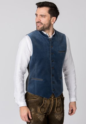 Blaues Samtwesten mit hohem Kragen, vorderem Knopfverschluss und zwei Taschen. Getragen über einem weißen Hemd mit Knopfdetails und dunkler Hose.