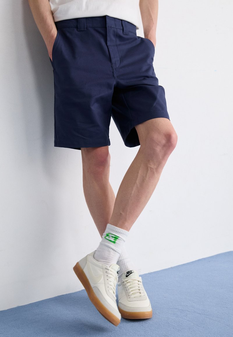 Pantaloni corti blu navy con vestibilità sartoriale, tasche laterali, abbinati a scarpe da ginnastica bianche e calzini con accenti verdi. Tessuto morbido e design pulito.