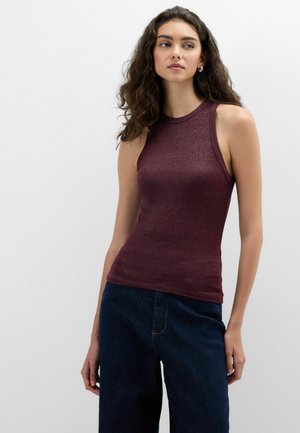 Vrouw met krullend haar die een mouwloos bordeaux rood topje en donkerblauwe spijkerbroek draagt, staand tegen een effen witte achtergrond.