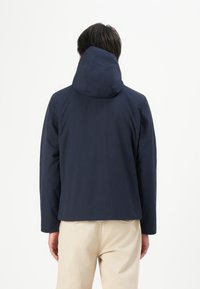 Man wearing a dark navy hooded jacket and beige pants, shown from the back against a plain white background.