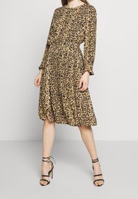 Vestido de estampado de leopardo en beige y negro, con mangas largas, falda plisada y cintura ajustada; combinado con tacones negros con tiras.