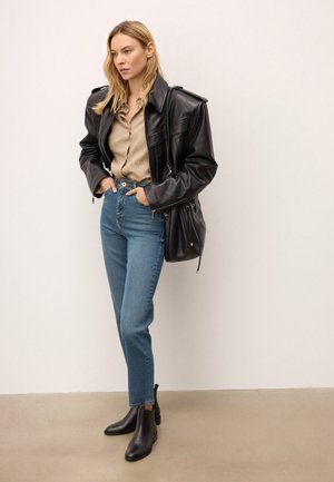Zwarte leren jas met schouderaccenten, beige blouse, hooghangende blauwe jeans en zwarte enkellaarsjes. Eenvoudige leren tas aan de zijkant.