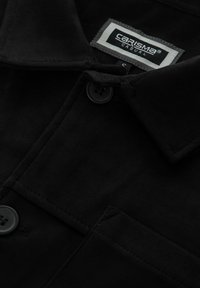 Camicia nera in materiale scamosciato con chiusura a bottoni, dotata di colletto, due tasche frontali e un'etichetta che indica "CARISMA CASUAL."