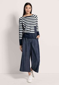 Frau in einem navy- und weißgestreiften langärmligen Oberteil mit aufgesetzter Tasche, weitgeschnittenen dunklen Denim-Culottes und weißen Sneakers, stehend.