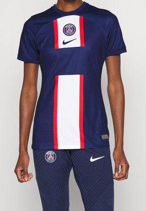 Person trägt ein marineblaues Paris Saint-Germain Fußballtrikot mit roten und weißen vertikalen Streifen und passende marineblaue Nike-Hose.