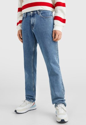 Personne portant un jean en denim bleu, un pull blanc à rayures rouges et des baskets blanches, debout devant un fond uni clair.