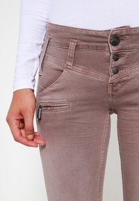 Jeans de mezclilla de color rosa claro con cintura alta, cierre de cuatro botones y un bolsillo con cremallera lateral. Textura suave con detalles de desvanecimiento sutil.