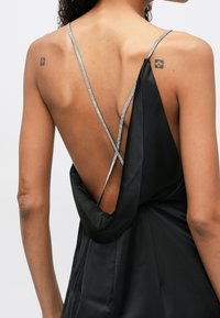 Femme portant une robe noire dos nu avec de fines bretelles croisées en strass et de petits tatouages visibles sur les deux épaules.