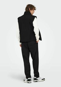 Schwarze ärmellose Weste über einem weißen Langarmshirt, ergänzt durch schwarze Hosen und schwarz-weiße Sneaker. Einfaches Design, keine Muster.