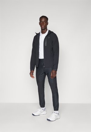 Sort letvægtsjakke med lynlås, hvid polo-shirt og mørke bukser. Strukturerede detaljer på jakken; sporty hvide sko.