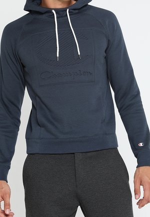 Muž v tmavě modré mikině Champion s bílými šňůrkami na kapuci a tmavě šedých teplácích, stojící s uvolněnýma rukama.