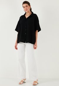 Black, oversized button-up shirt with short sleeves, textured fabric. Paired with white, wide-leg pants and strappy sandals.
