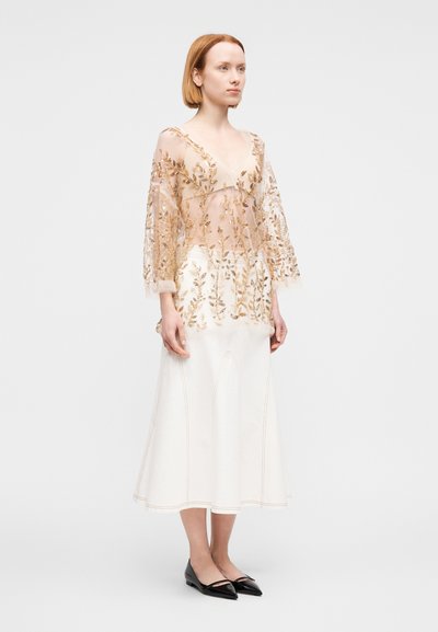 Διάφανο, μπεζ τοπ με χρυσή κεντητή ανθοδέσμη, σε συνδυασμό με φουσκωτή λευκή φούστα με ραφές και μαύρες μυτερές μπαλαρίνες.