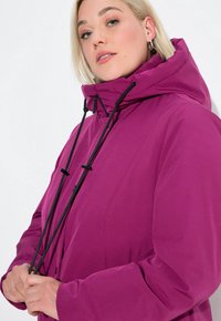 Blonde woman adjusting black drawstrings on a magenta hooded jacket against a white background.