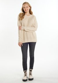 Pulover bej din tricot cu model cordonat, cu tiv ondulat, purtat cu blugi skinny negri și botine bej cu șireturi. Fundalul este alb simplu.