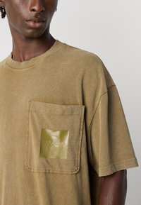 Mand iført en falmet olivengrøn T-shirt med en stor brystlomme, der har et glansfuldt "HUGO" logo.