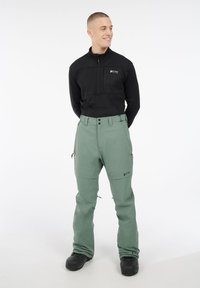 Zwarte fleece trui met ritssluiting, gecombineerd met groene waterdichte broek. De broek heeft zakken en een getailleerde pasvorm. Model draagt zwarte schoenen.