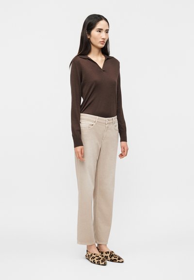 Femme debout portant une chemise marron à manches longues, un pantalon beige taille haute et des chaussures slip-on à motif léopard, sur un fond blanc uni.