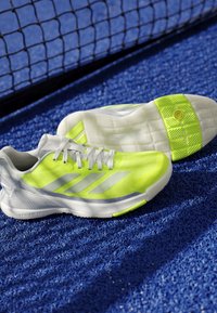 White and neon yellow tennis shoes on a blue court surface near a black tennis net.