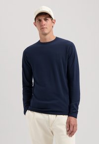 Navyblaues langärmliges T-Shirt aus weichem Stoff, mit Rundhalsausschnitt und dezentem Logo auf der Brust. Kombiniert mit cremefarbenen Hosen.