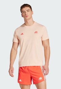 Světle růžové tričko s krátkými rukávy, na kterém je červené logo a červené pruhy. Kombinováno s oranžovými šortkami, které mají minimalistický design.